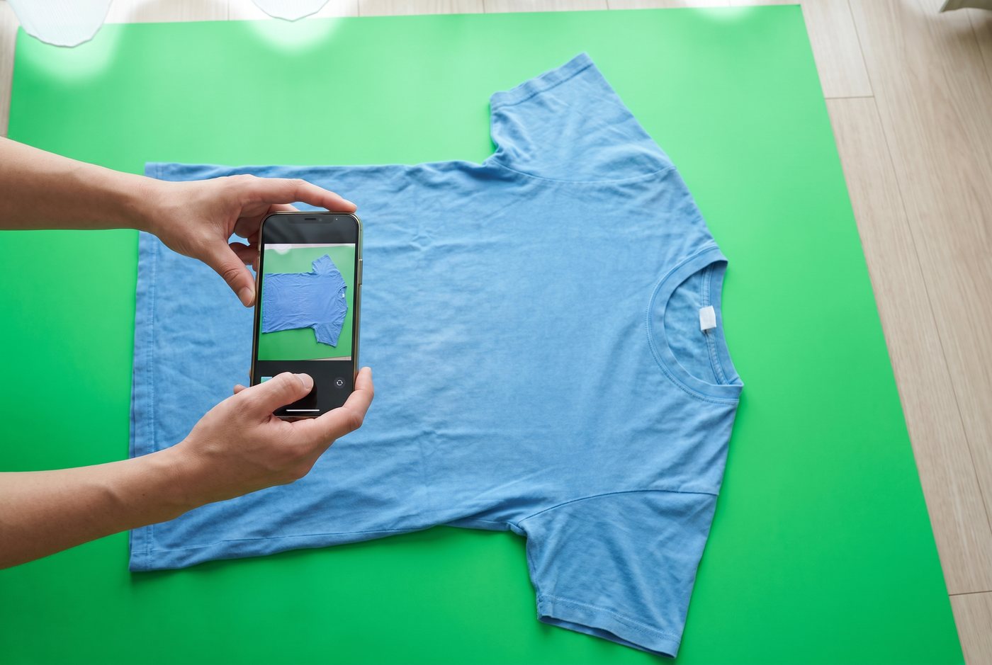 Deux mains photographient au téléphone un t-shirt posé à plat sur un drap vert